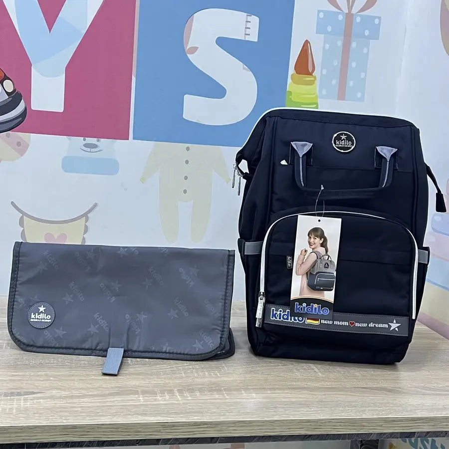 Kidilo dark diaper backpack and grey printed changing mat on a wooden surface, with a product tag on the front.