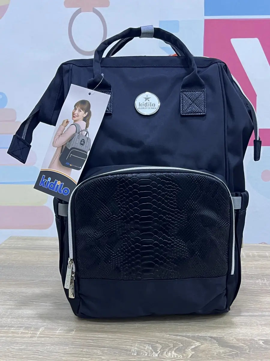 Kidilo blue diaper backpack on a wooden surface, front view with textured pocket, round badge and hanging tag.