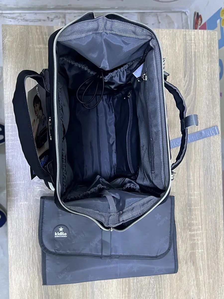 Kidilo dark diaper backpack open on a wooden surface, with grey-lined inner pockets, cord and changing mat.